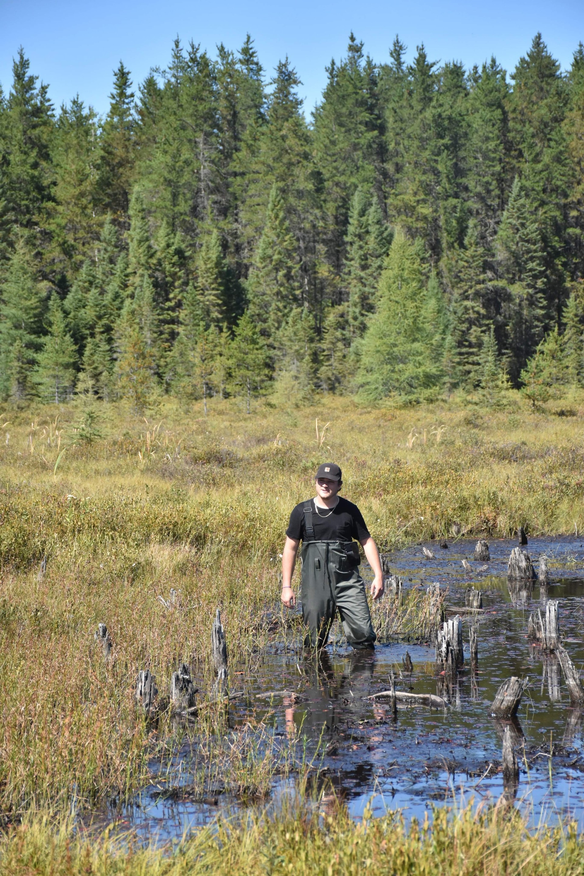 Frédérick Girard, étudiant à la maîtrise en sciences de l’environnement (profil avec mémoire) de l’UQTR, porte une salopette imperméable lors d'une sortie sur le terrain. Conifères à l'arrière, végétation ambiante et de l'eau jusqu'aux genoux.