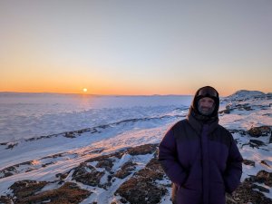 Laurent Lessard est étudiant à la maîtrise en sciences de l’environnement (profil avec mémoire) de l’Université du Québec à Trois-Rivières (UQTR). Il est ici photographié dans le Grand Nord, habillé avec des vêtements chauds, devant de vastes étendues de neige et de roche, avec un soleil bas donnant une teinte orangée au paysage.