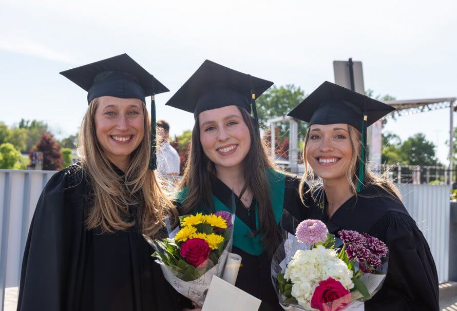 Trois étudiantes portant une toge et un mortier participent à leur cérémonie de collation des grades. Elles tiennent deux bouquets de fleurs et un parchemin de diplôme.