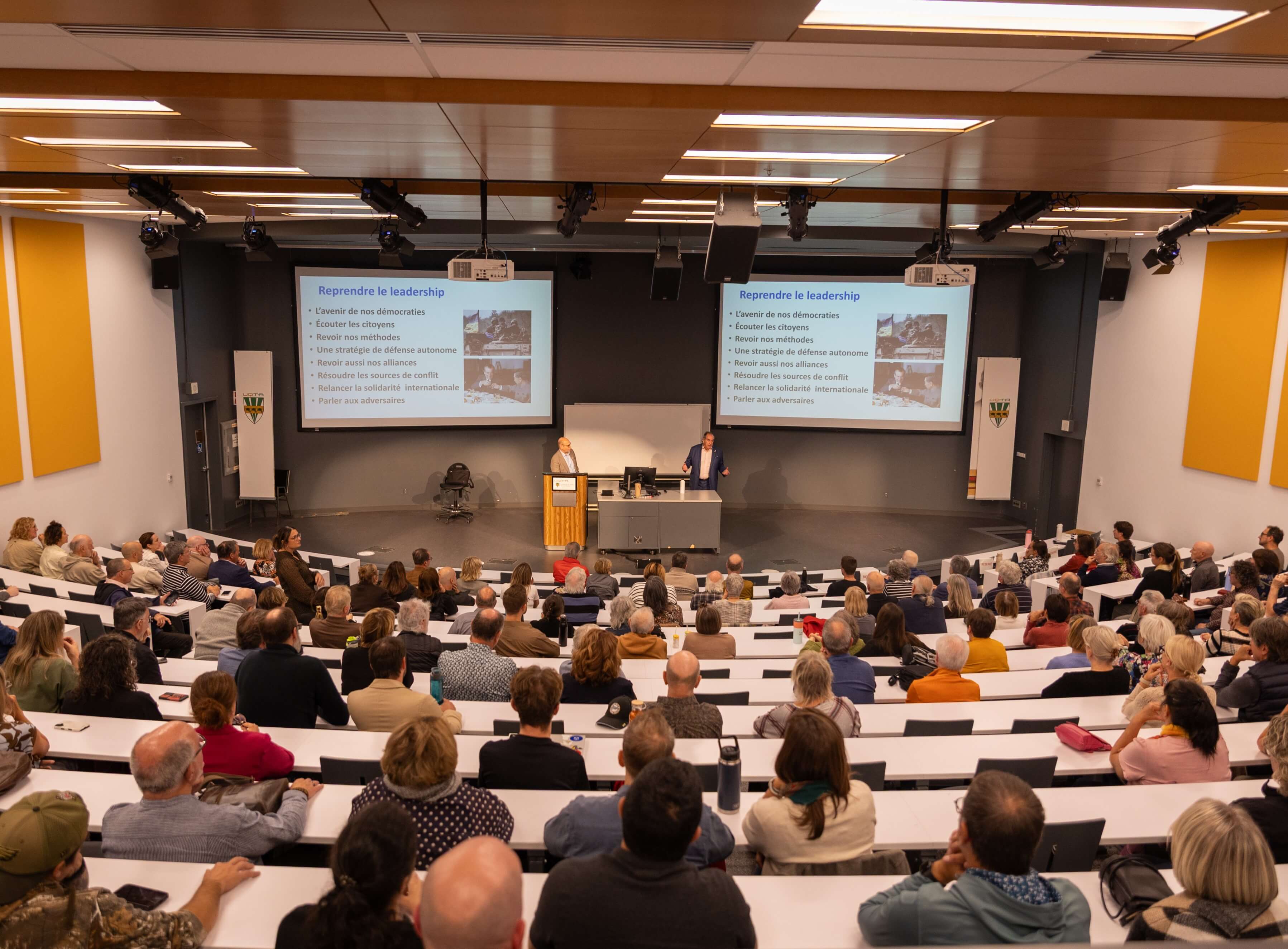 Un auditorium bondé, avec Jean-François Lépine faisant une présentation à l'avant.