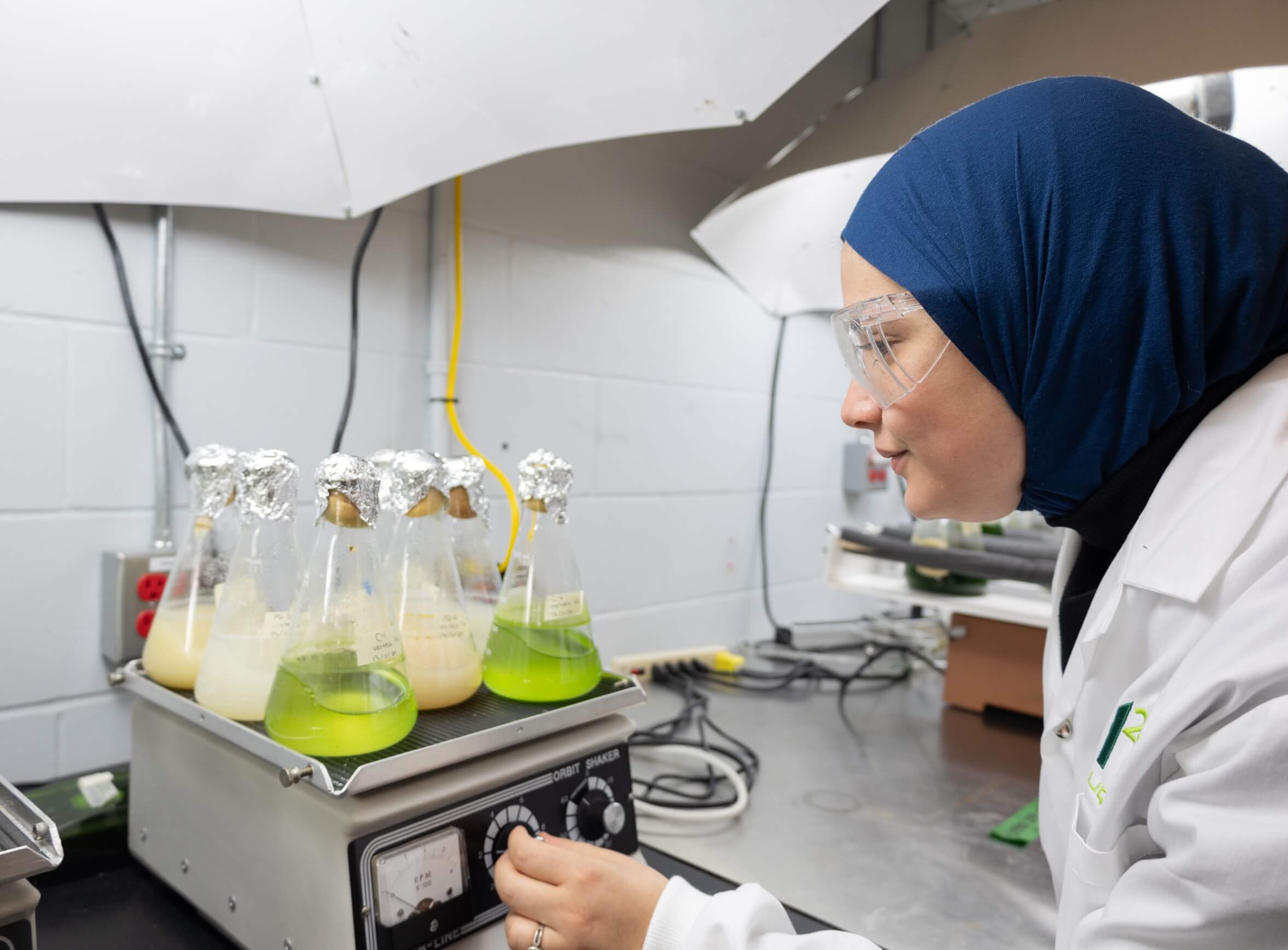 Une étudiante règle un appareil sur lequel sont déposés des erlenmeyers (fioles coniques) contenant un liquide vert ou trouble.